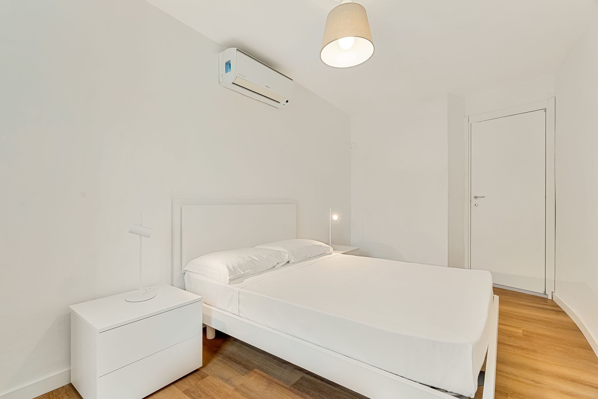A minimalist bedroom with a bed, bedside table, nightstand, and an air conditioning unit.