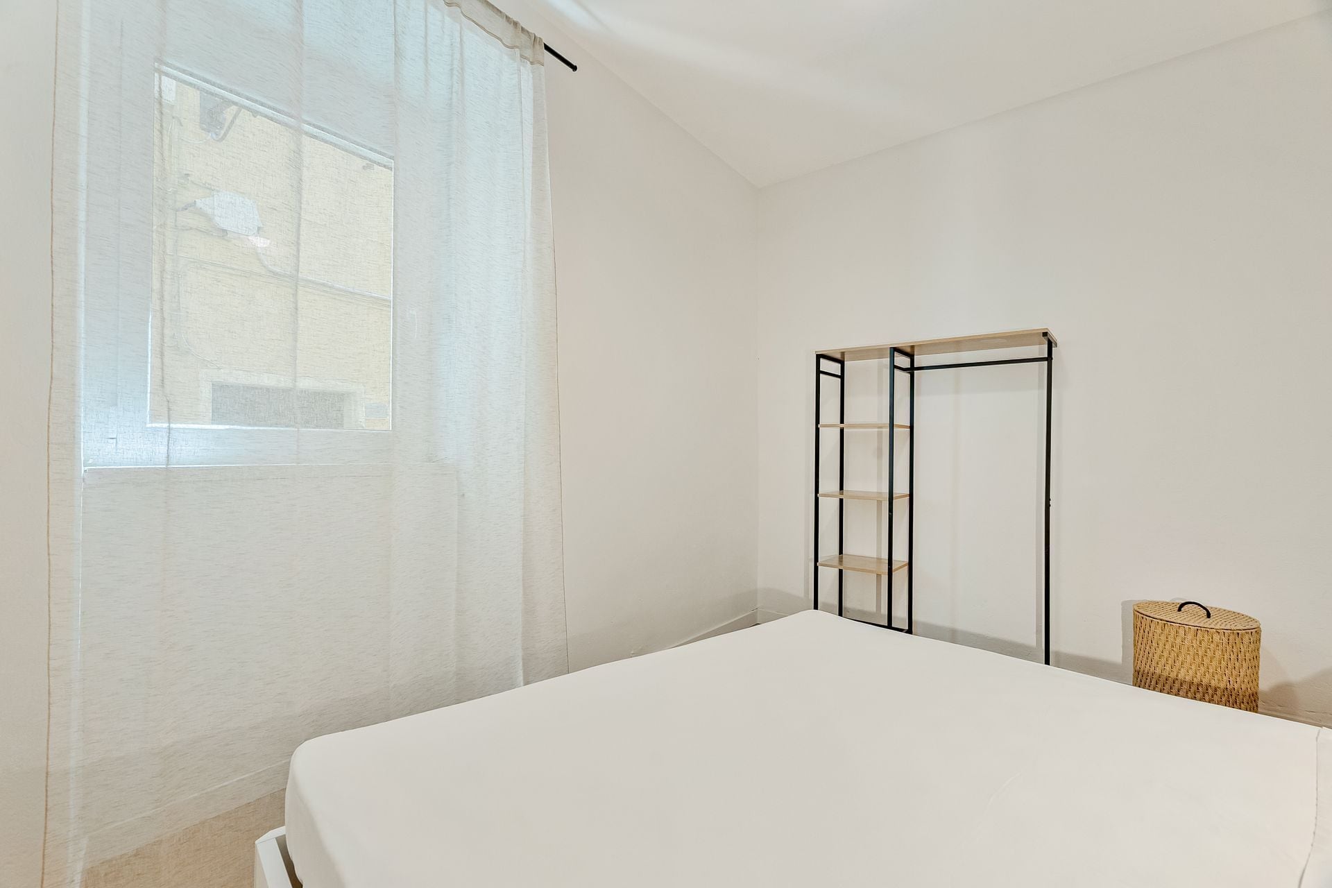 Una camera da letto con un letto, una finestra con tende leggere, un armadio in metallo e un cesto di vimini.
