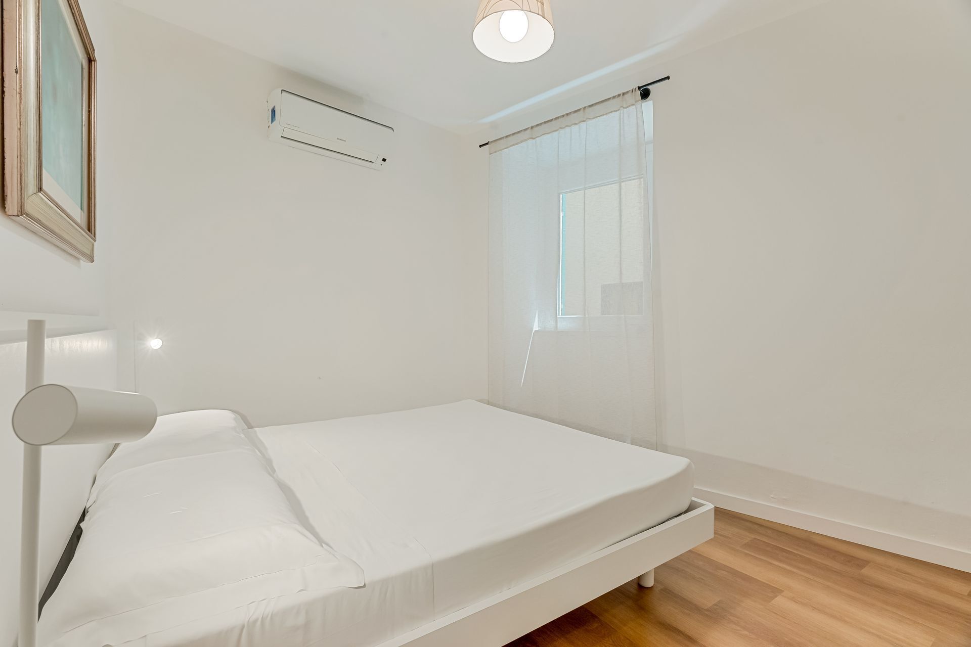 Une petite chambre avec un lit simple, un matelas blanc, une table de chevet, un luminaire au plafond, une fenêtre avec des rideaux légers et un climatiseur fixé au mur.