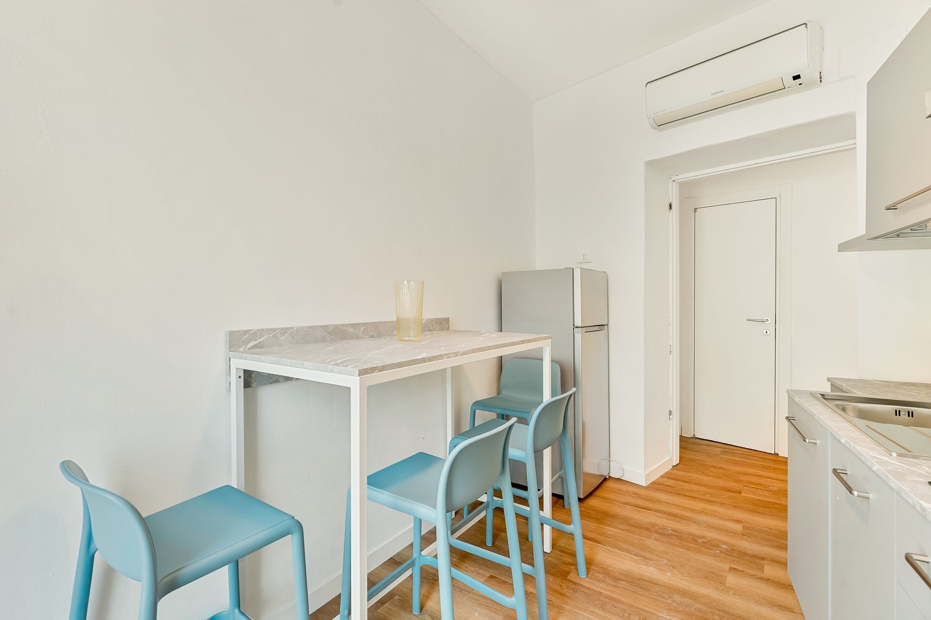A compact kitchen with a white countertop, a refrigerator, and two blue chairs.