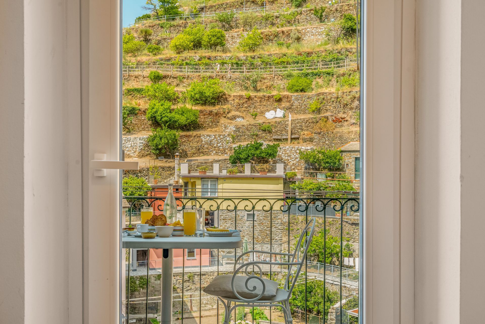 A balcony with a table set for breakfast, a chair, and a view of a hillside with buildings.