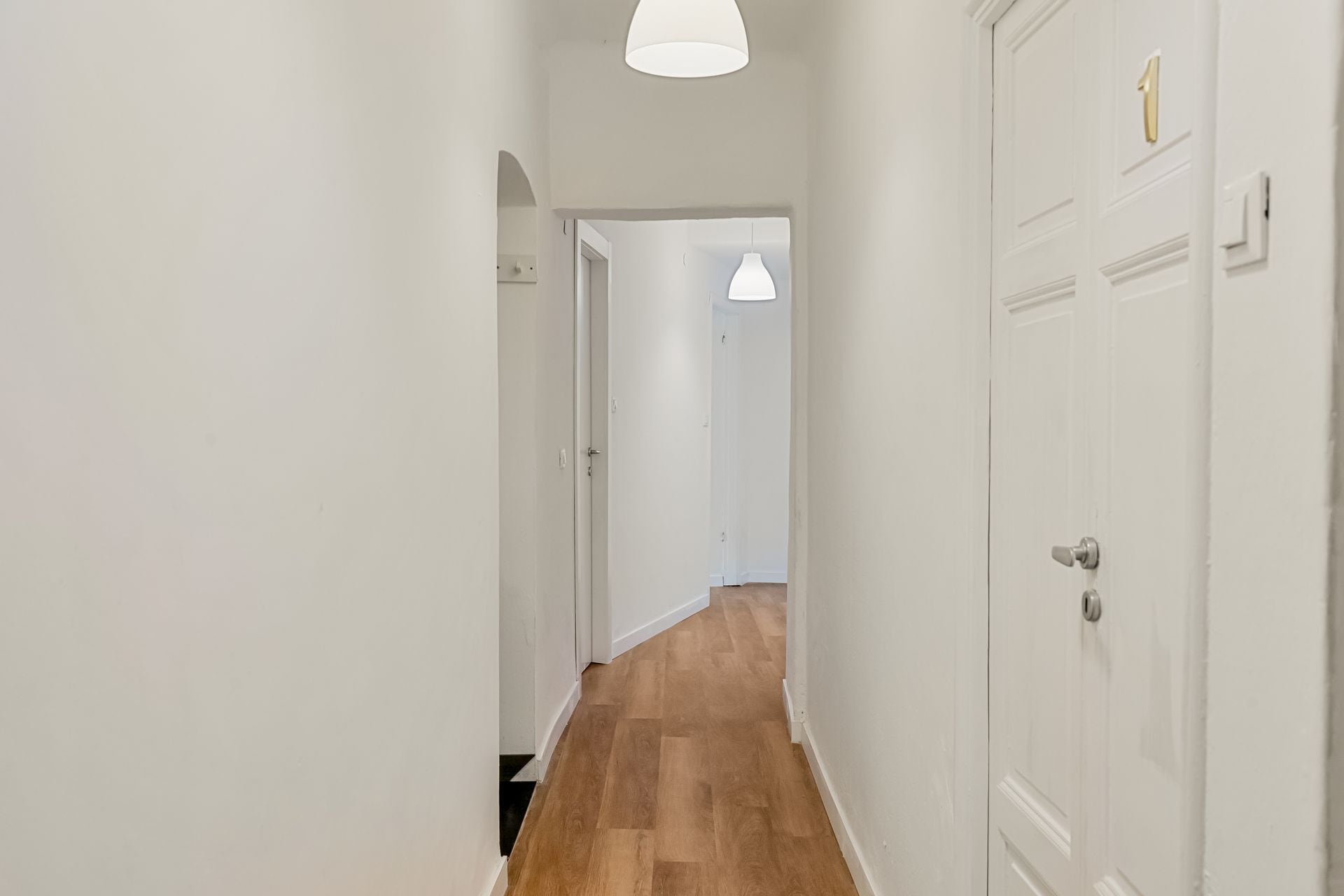 A hallway with wooden flooring, white walls, and doors with a number plate.