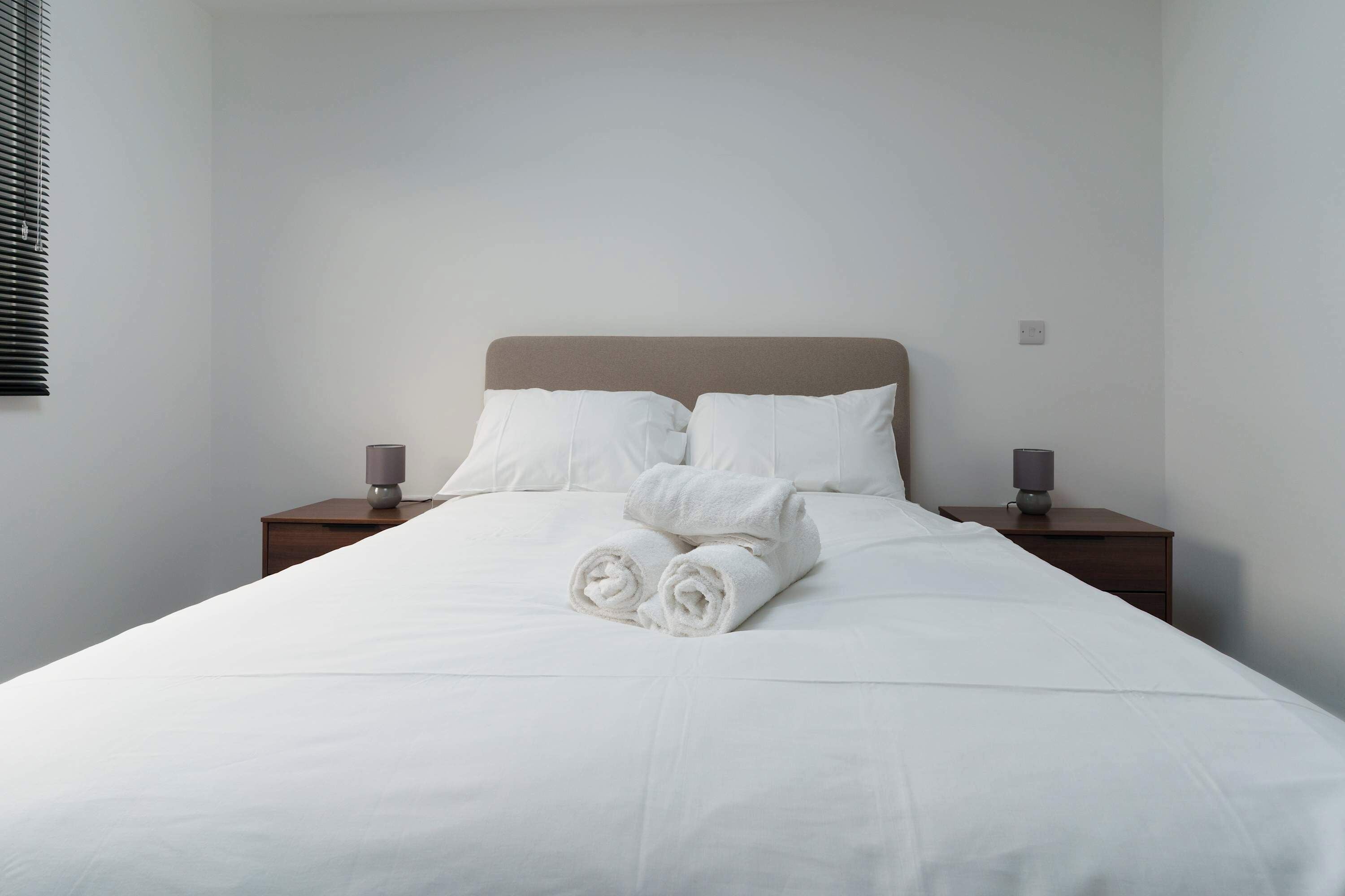 A neatly made bed with white linens and rolled towels, two bedside tables with lamps, and a plain wall.
