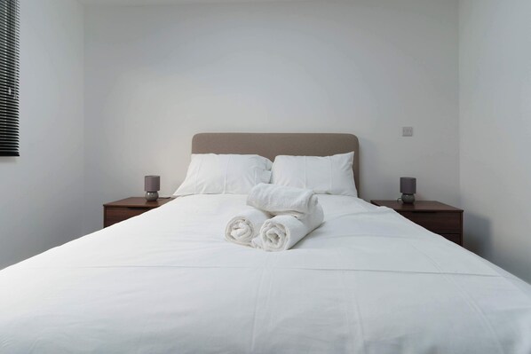 A neatly made bed with white linens and rolled towels, two bedside tables with lamps, and a plain wall.