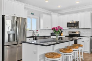 A modern kitchen with white cabinetry, stainless steel appliances, a black countertop island, and wicker stools. - Endless Summer, Oceanfront, 8 Bdrm, Pvt. Htd Pool, Pvt. Direct Beach Access (Holden Beach)