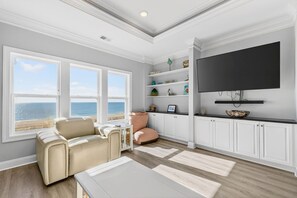 A living room with a sofa, a coffee table, a TV, and a view of the ocean through large windows. - Endless Summer, Oceanfront, 8 Bdrm, Pvt. Htd Pool, Pvt. Direct Beach Access (Holden Beach)