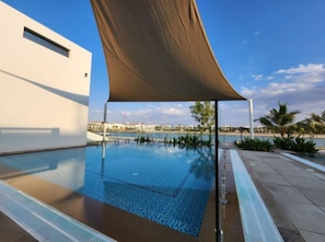 Piscina con vistas despejadas al mar, zona sombreada y un edificio moderno al fondo.