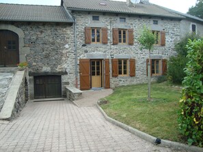 Un edificio de piedra con entrada empavedada, contraventanas de madera y un pequeño jardín.