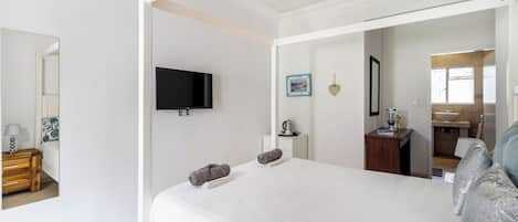 A bedroom with a four-poster bed, a flat-screen TV mounted on the wall, a bedside table with a lamp, and a bathroom visible through an open door.