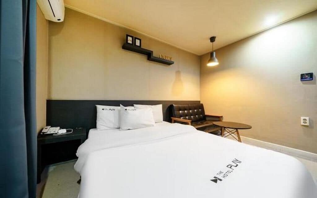 A hotel room with a bed, a bedside table with a phone, a small table, a chair, and a wall-mounted shelf with a lamp.