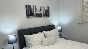 Ein Schlafzimmer mit einem Bett, zwei Nachttischen mit Lampen, einem an der Wand hängenden Stadtbild und einem Fenster mit Jalousien.
