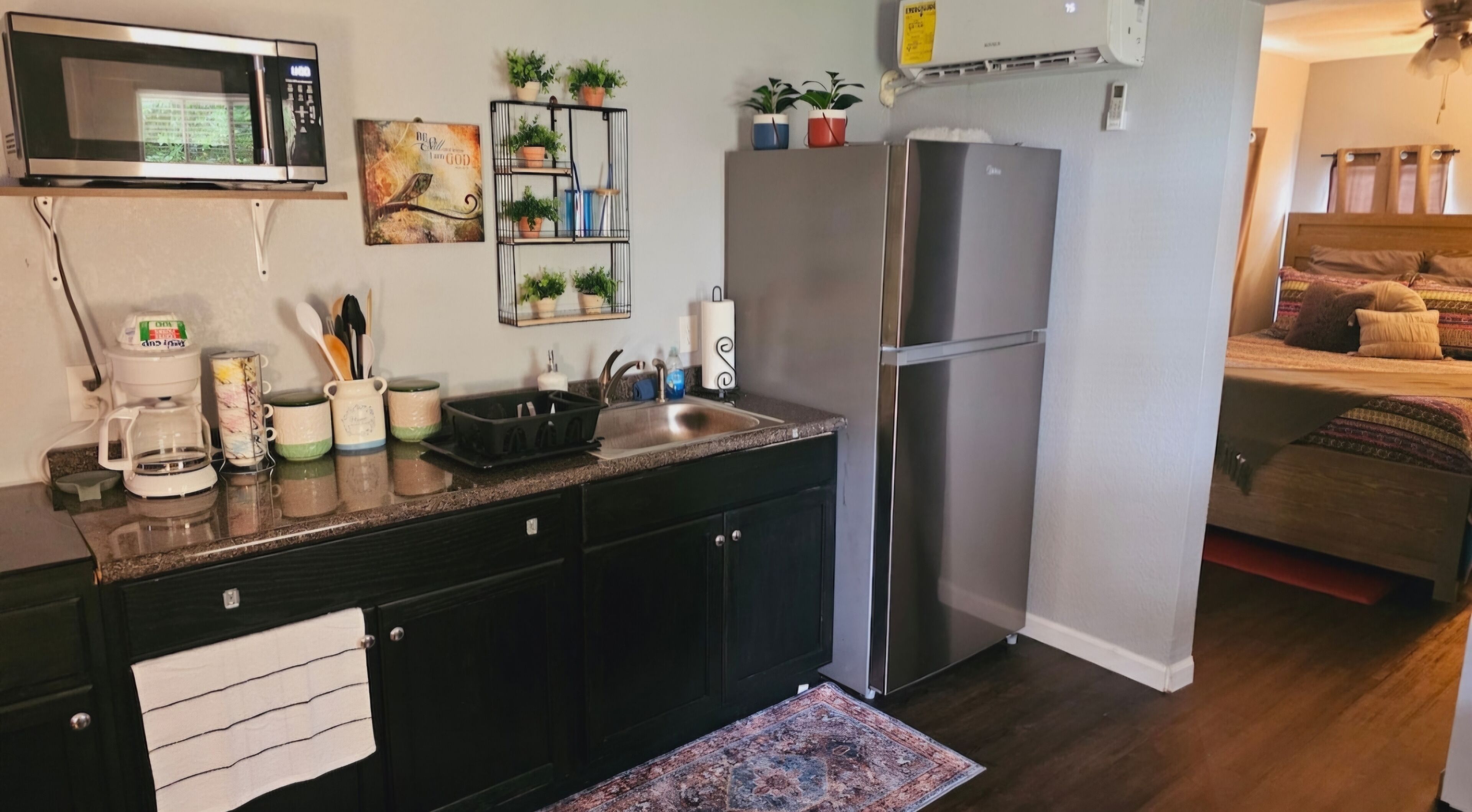 A compact kitchen with a microwave, coffee maker, and a refrigerator. There are potted plants on a shelf and a rug on the floor.