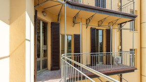 Ein Balkon mit Metallgeländer, ein Fenster mit Rollo und eine Tür zu einem Innenraum.