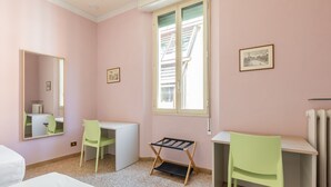 Ein Zimmer mit zwei Schreibtischen, zwei Stühlen, einem Bett, einer Heizung und einem Fenster mit Blick auf Gebäude.