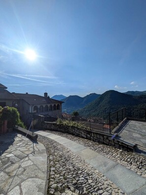 A stone pathway leading to a building with a tiled roof, surrounded by mountains and a clear sky. - Casa Panorama — 50 square meters of garden with a hot tub and a view of the lake (Laino)