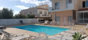 A swimming pool with lounge chairs, a white fence, and a building with balconies. - Venus Beach Apartment. Diana 33 (Pafos)