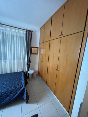 A bedroom with a bed, a wardrobe, a small stool, and a window with curtains. - Venus Beach Apartment. Diana 33 (Pafos)
