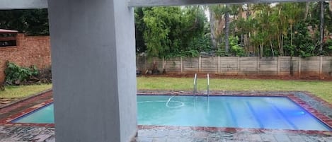 A covered outdoor swimming pool with a tiled deck, a metal ladder, and a brick wall in the background.