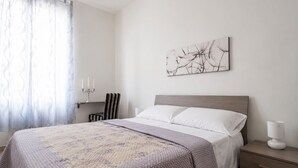 A bedroom with a bed, a bedside table, a lamp, a chair, a wall-mounted artwork, and a window with curtains.