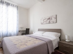 A bedroom with a bed, a bedside table, a lamp, a chair, a wall-mounted artwork, and a window with curtains. - Elegant apartment just a stone's throw from Piazza del Campo (Siena)