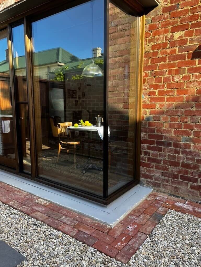 Une porte vitrée donne sur un patio aménagé avec une table et des chaises, un mur en briques et un sol en gravier.