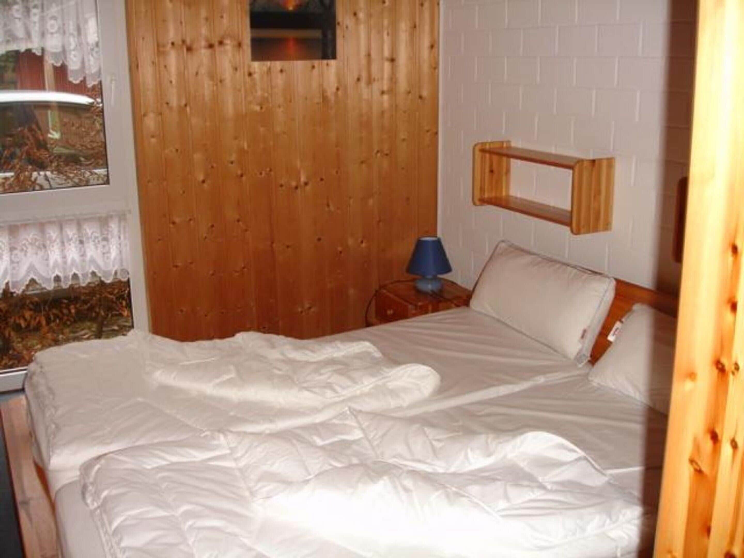Een slaapkamer met een houten hoofdeinde, een bed met witte beddengoed, een nachtkastje met een lamp en een raam met gordijnen.
