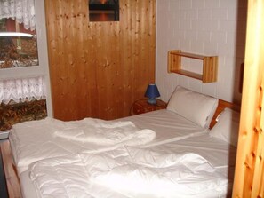 Ein Schlafzimmer mit Holzkopfteil, einem Bett mit weißer Bettwäsche, einem Nachttisch mit Lampe und einem Fenster mit Vorhängen.