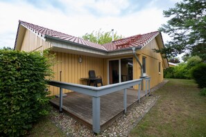 Ein eingeschossiges Holzhaus mit Veranda, einer kleinen Terrasse und einem überdachten Sitzbereich im Freien.