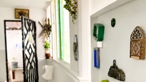 En hall med ett dekorativt metallgaller, en krukväxt, ett fönster med gröna fönsterluckor och en vägg med diverse föremål, inklusive en grön flaska, en blå behållare och en dekorativ låda.