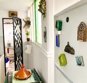 Ein Flur mit einem verzierten Metallgitter, einem Blumentopf, einem Fenster mit grünen Rollläden und einer Wand mit verschiedenen Gegenständen, darunter eine grüne Flasche, ein blauer Behälter und eine Dekobox.