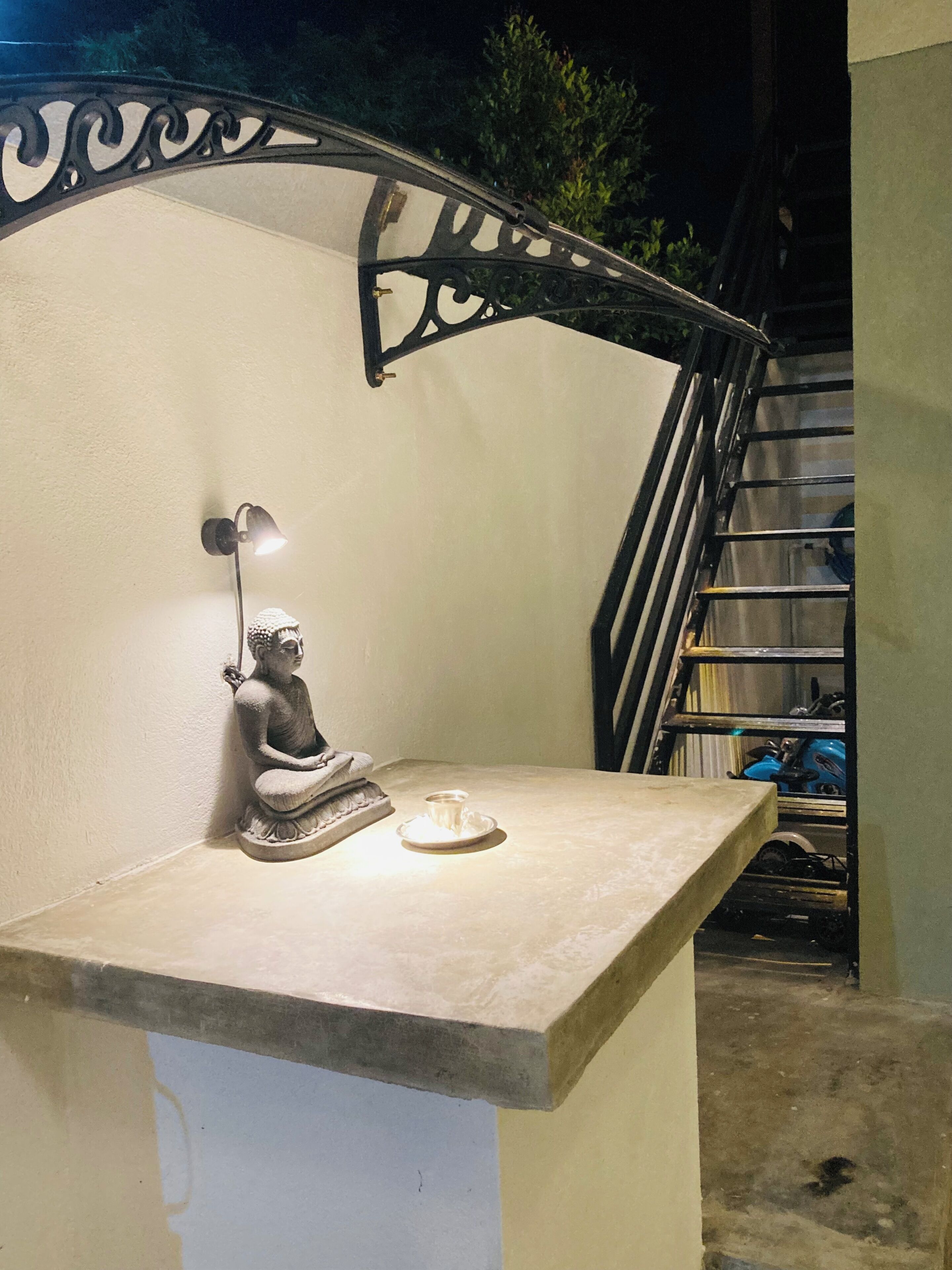 A statue of a seated figure on a table with a lamp and a cup, against a wall with a metal railing.