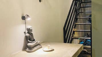 A statue of a seated figure on a table with a lamp and a cup, against a wall with a metal railing.