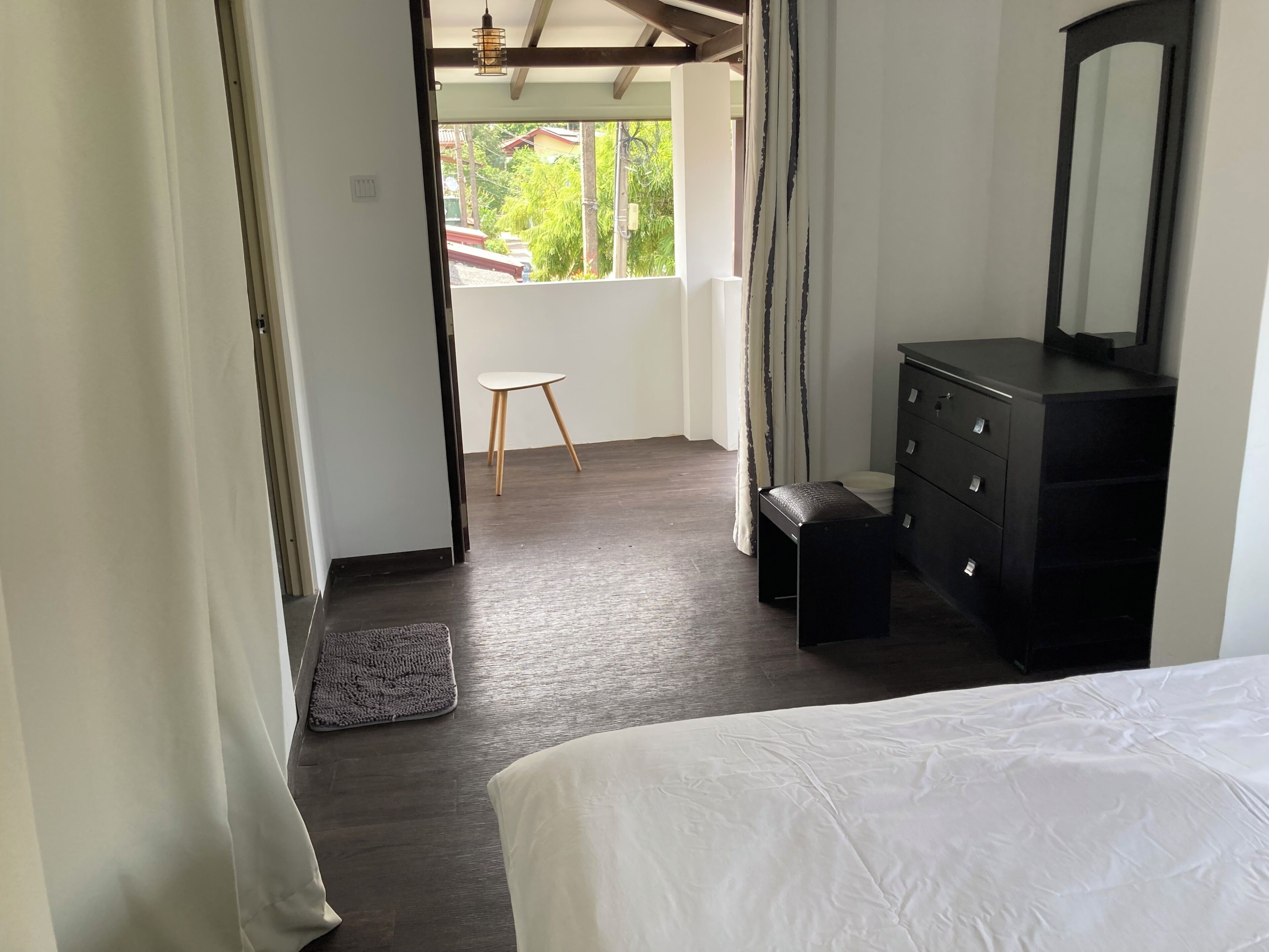 A bedroom with a bed, a dresser, a mirror, a small stool, and a view of the outdoors.