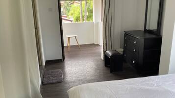A bedroom with a bed, a dresser, a mirror, a small stool, and a view of the outdoors.
