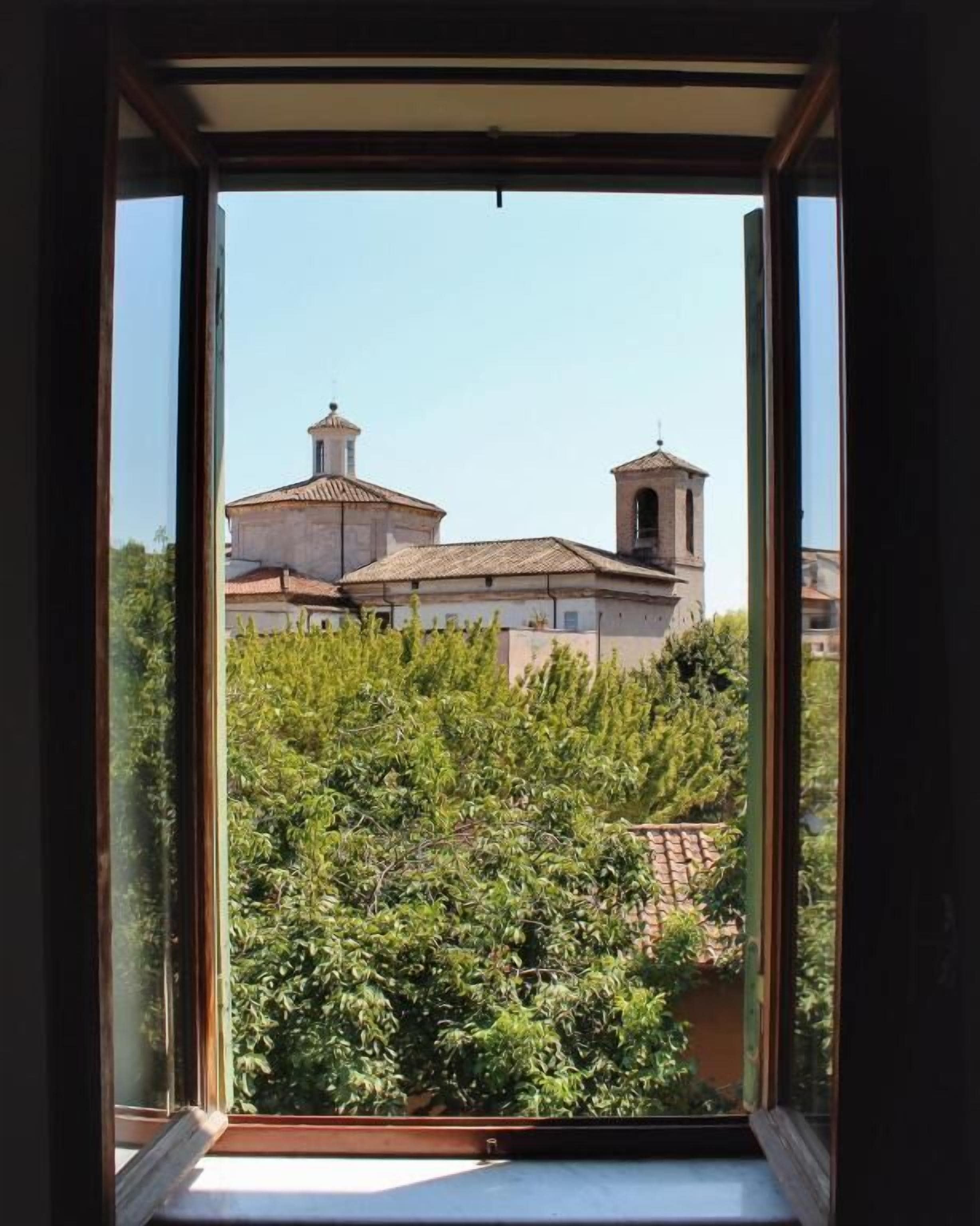Visão pela janela de um edifício com uma torre de sino e uma cúpula, rodeado de vegetação.