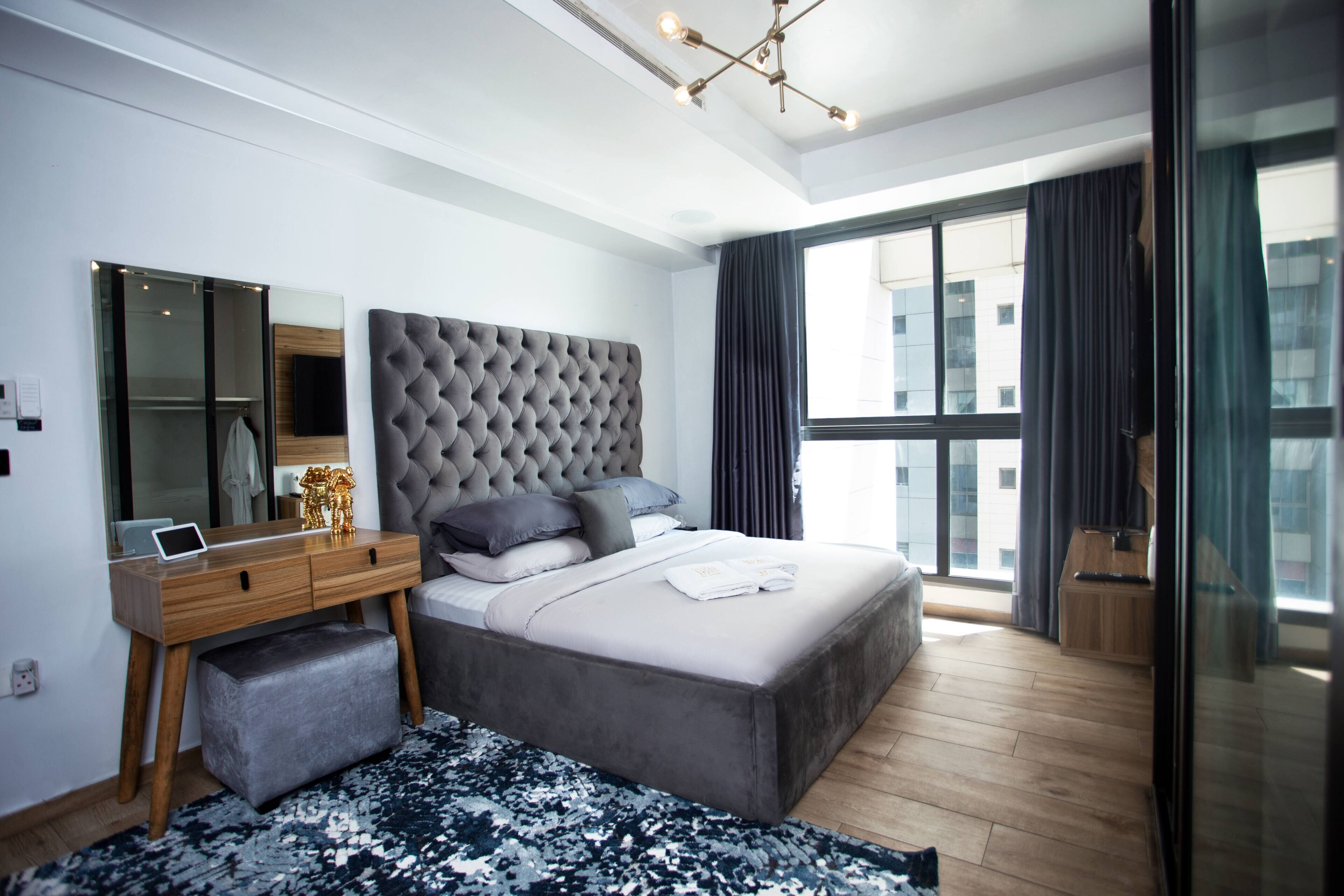 A modern bedroom with a large bed, a wooden dresser, a mirror, and a large window with curtains.