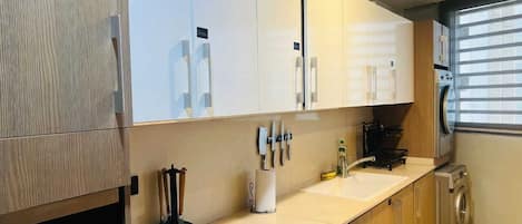 A modern kitchen with wooden cabinets, a countertop, a sink, a washing machine, and a window with blinds.