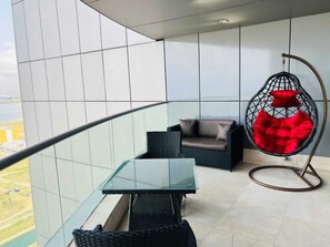 A balcony with a glass table, wicker furniture, and a red hanging chair. - Eko Atlantic Apartment (Lagos)
