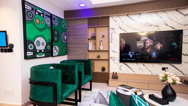 A modern living room with a flat-screen TV, green velvet chairs, a marble wall, and a shelf with decorative items. - Eko Atlantic Apartment (Lagos)