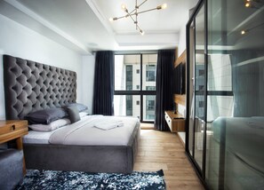 A modern hotel room with a large bed, a wooden nightstand, a built-in bench, a large window with curtains, and a ceiling light fixture. - Eko Atlantic Apartment (Lagos)