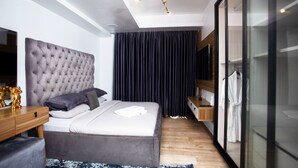 A modern bedroom with a tufted headboard, a wooden bedside table, a bedside lamp, a large window with curtains, and a built-in wardrobe. - Eko Atlantic Apartment (Lagos)