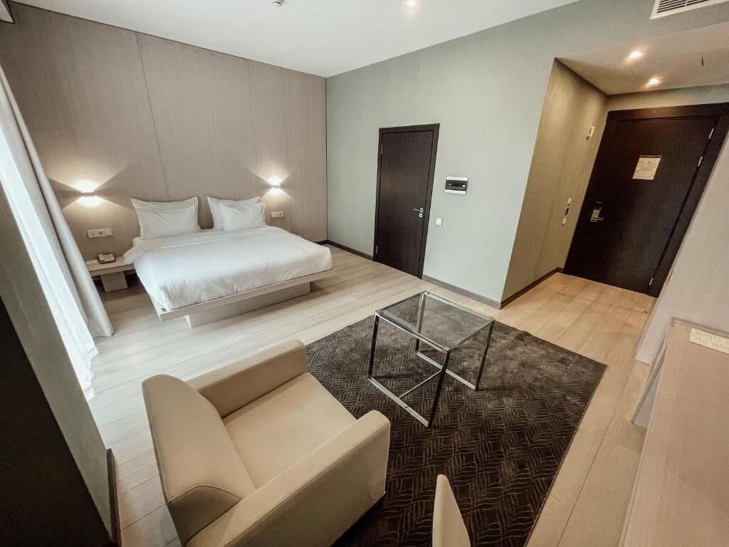 Una habitación de hotel moderna con una cama, mesitas de noche, un sofá, una mesa de centro de vidrio y una alfombra estampada.