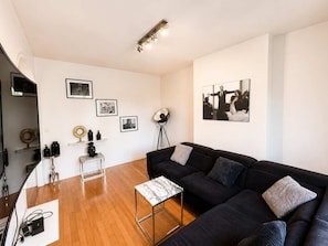 Een moderne woonkamer met een zwarte bank, een klein bijzettafeltje en een vloerlamp. Er hangen afbeeldingen in lijsten aan de muur en een groot raam zorgt voor natuurlijk licht.