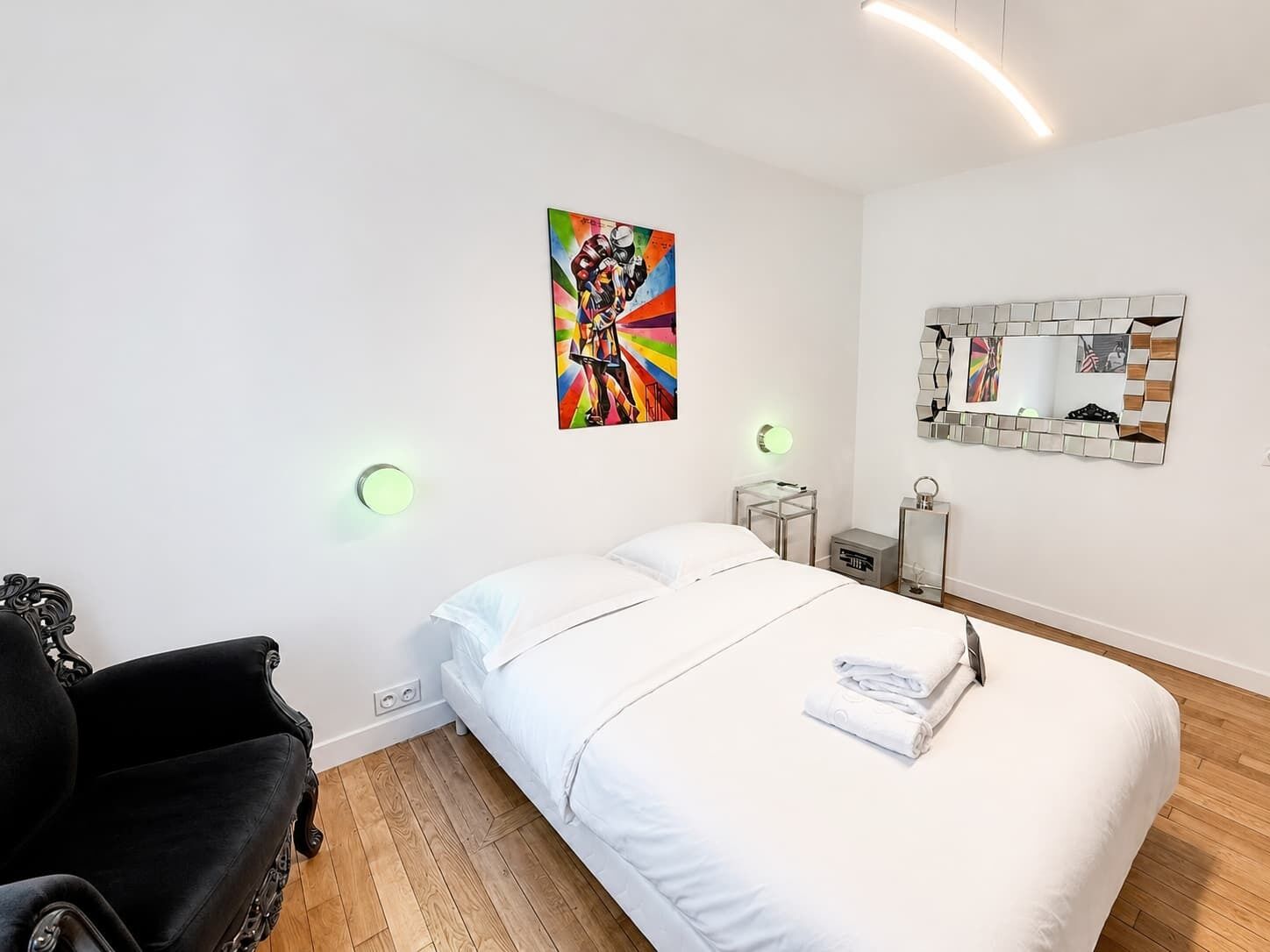 A hotel room with a bed, a chair, a painting, a mirror, and bedside tables.