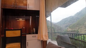 Una habitación con un armario de madera, una silla, una mesita con un hervidor y un balcón con vistas a un paisaje montañoso.