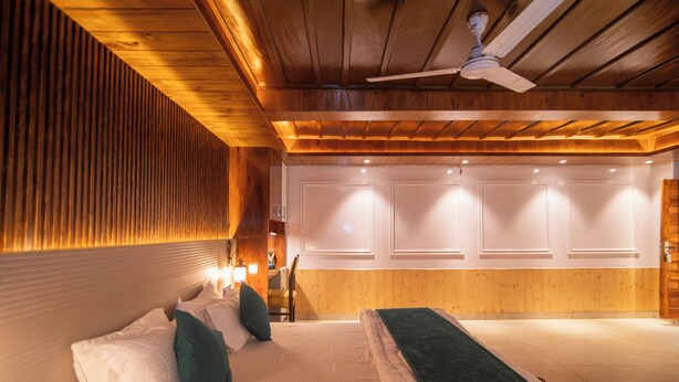 A hotel room with a wooden ceiling, a bed with pillows, a bedside table, and a wall-mounted light.