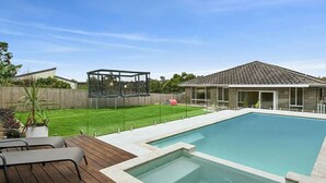 Ein Schwimmbad mit Holzterrasse, einem überdachten Spielbereich und einem Haus im Hintergrund.