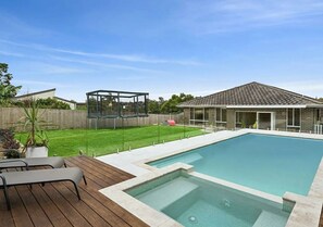 Ein Schwimmbad mit Holzterrasse, einem überdachten Spielbereich und einem Haus im Hintergrund.