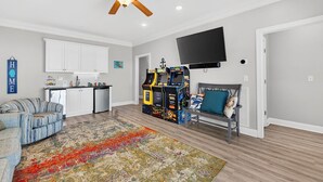 Een woonkamer met een bank, een salontafel, een kleurrijk vloerkleed, een plafondventilator, een flatscreen-tv en een rij arcadekastjes.