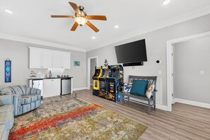 A living room with a sofa, a coffee table, a colorful rug, a ceiling fan, a flat-screen TV, and a row of arcade games. - Endless Summe, Oceanfront 8 Bedroom w/ Heated Pool (Holden Beach)